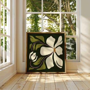 May include: Framed art print depicting a stylized white magnolia flower with a yellow centre and purple stems against a dark green backdrop. The artwork is displayed in a room with natural light from a window.