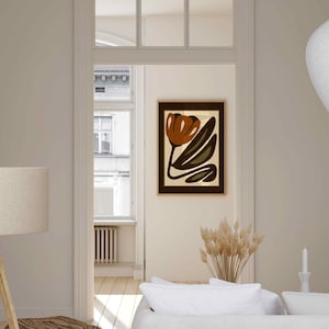 K&ouml;nnte beinhalten: Ein abstrakter Kunstdruck mit einem braunen Tulpendesign mit gr&uuml;nen Bl&auml;ttern auf cremefarbenem Hintergrund, gerahmt in einem dunkelbraunen Holzrahmen. Das Kunstwerk h&auml;ngt an einer cremefarbenen Wand in einem Raum mit einem wei&szlig;en Sofa und einer Stehlampe.
