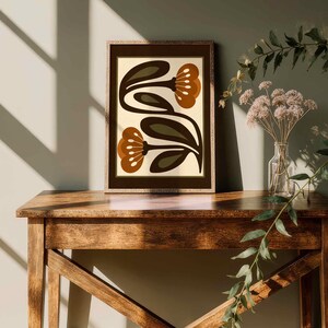 Puede incluir: Una impresi&oacute;n art&iacute;stica enmarcada con un dise&ntilde;o floral retro en tonos marr&oacute;n, verde oliva y crema. La obra de arte se exhibe sobre una mesa de madera. La escena se realza con luz natural y sombras. Tambi&eacute;n hay un jarr&oacute;n con flores secas y vegetaci&oacute;n.