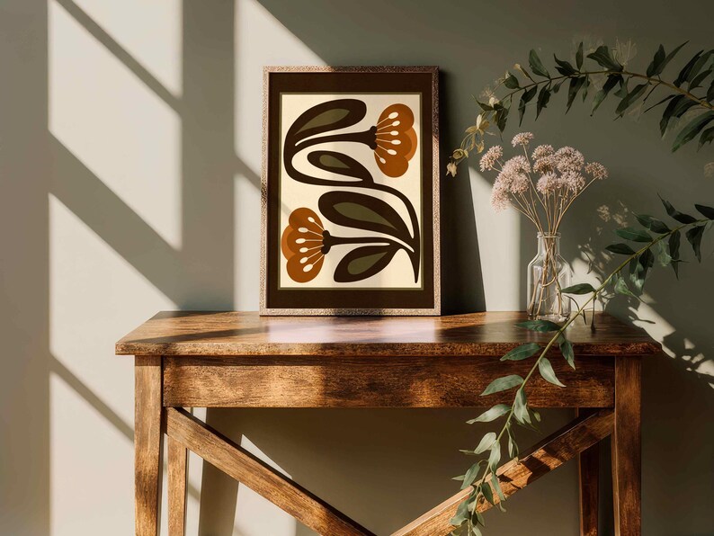 Puede incluir: Una impresi&oacute;n art&iacute;stica enmarcada con un dise&ntilde;o floral retro en tonos marr&oacute;n, verde oliva y crema. La obra de arte se exhibe sobre una mesa de madera. La escena se realza con luz natural y sombras. Tambi&eacute;n hay un jarr&oacute;n con flores secas y vegetaci&oacute;n.