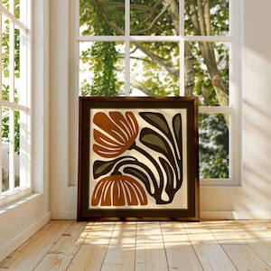 May include: A framed art print featuring a stylized floral design. The artwork showcases two large, rust-colored flowers with cream-colored petals and dark brown and olive green leaves against a cream background. The frame is dark brown.
