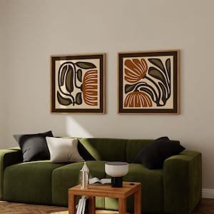 May include: Two framed floral art prints with brown, olive green, and cream-colored abstract designs. The prints are displayed above a green sofa with black and white pillows. The room has a modern, minimalist aesthetic.
