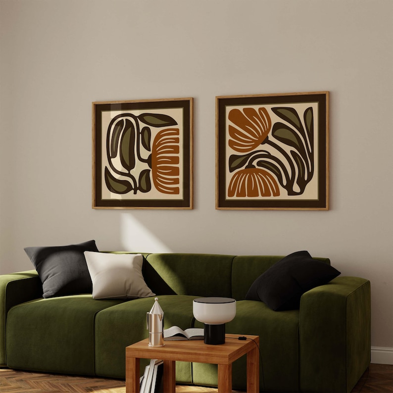 May include: Two framed floral art prints with brown, olive green, and cream-colored abstract designs. The prints are displayed above a green sofa with black and white pillows. The room has a modern, minimalist aesthetic.