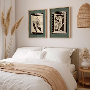 Op de afbeelding: Twee ingelijste botanische kunstprints met een groene rand en een natuurlijke houten lijst hangen boven een bed. De prints tonen zwarte en cr&egrave;mekleurige bloemmotieven. Het bed is opgemaakt met wit beddengoed en een bruine sprei.