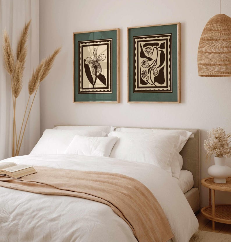 Op de afbeelding: Twee ingelijste botanische kunstprints met een groene rand en een natuurlijke houten lijst hangen boven een bed. De prints tonen zwarte en cr&egrave;mekleurige bloemmotieven. Het bed is opgemaakt met wit beddengoed en een bruine sprei.