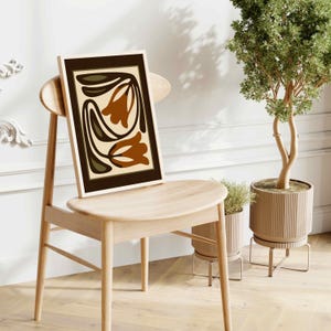 May include: A framed art print featuring a floral design in shades of brown, olive green, and cream. The artwork is displayed on a light wood chair, with a potted plant in the background. The print has a dark brown border.