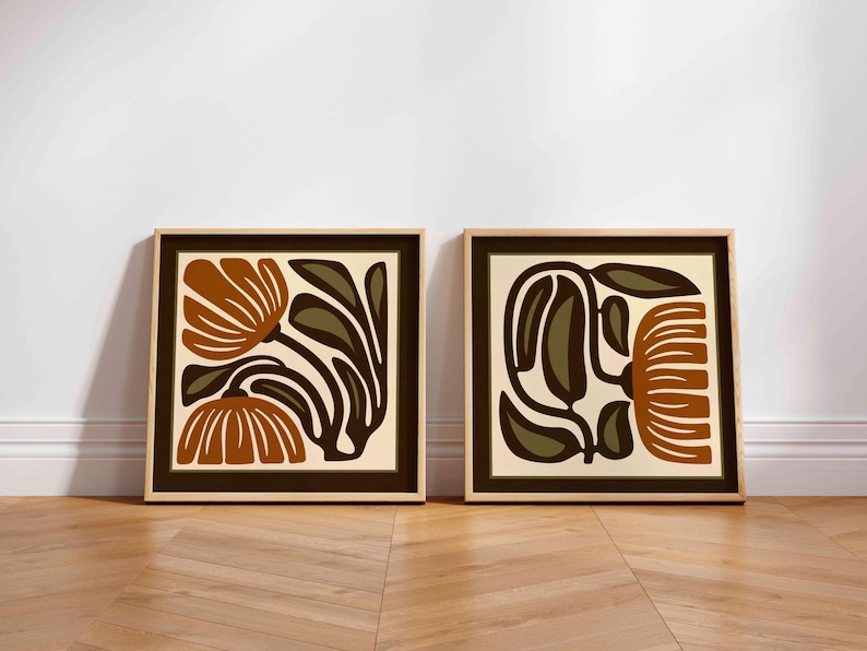 May include: Two framed floral art prints. Each print features a stylized floral design in shades of brown, olive green, and cream. The frames are a light wood color, and the artwork is displayed against a white wall.