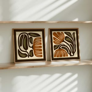 May include: Two framed art prints with abstract floral designs. Each print features a beige background with brown and rust-coloured floral shapes and dark green leaves. The frames are a dark brown colour, and the prints are displayed on a wooden shelf.
