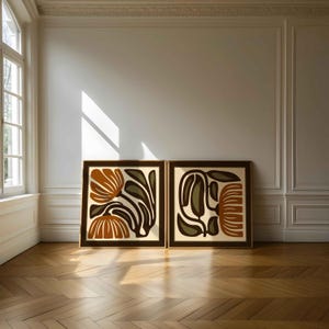 May include: Two framed art prints with abstract floral designs. The artwork features orange, brown, and olive green shapes on a cream background. The frames are dark brown, and the prints are displayed on a wooden floor.
