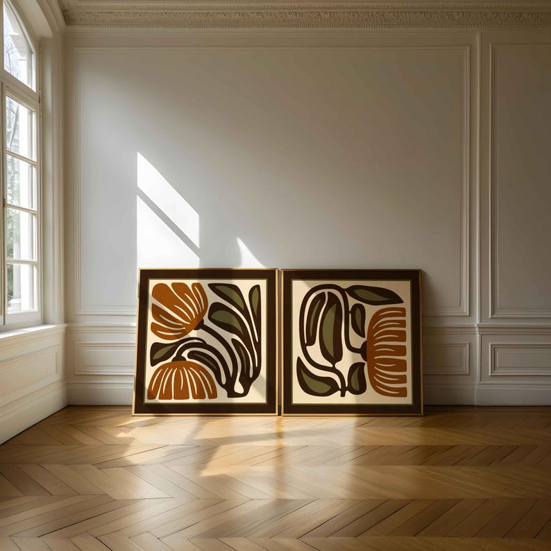 May include: Two framed art prints with abstract floral designs. The artwork features orange, brown, and olive green shapes on a cream background. The frames are dark brown, and the prints are displayed on a wooden floor.
