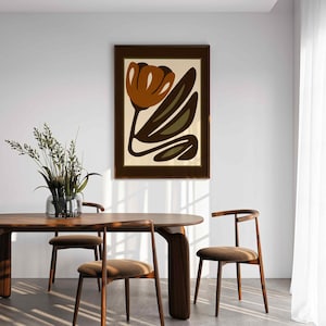 K&ouml;nnte beinhalten: Ein gerahmter Kunstdruck mit einer stilisierten Blume in Braun- und Olivgr&uuml;nt&ouml;nen auf cremefarbenem Hintergrund. Das Kunstwerk h&auml;ngt an einer wei&szlig;en Wand &uuml;ber einem Esstisch und St&uuml;hlen aus Holz und schafft ein modernes Interieur.