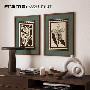 Op de afbeelding: Twee ingelijste botanische prenten met walnootframes en groene passe-partouts. De prenten tonen zwarte en cr&egrave;mekleurige bloemmotieven. Een donkerbruine tafellamp, kaarsen en decoratieve objecten zijn gerangschikt op een donkerbruine kast. De tekst "Frame: Walnut" staat bovenaan.