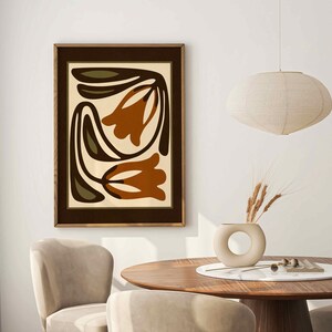 May include: A framed abstract art print featuring stylized orange and brown tulip-like shapes on a cream background. The artwork is in a dark brown frame, displayed above a wooden table with chairs. A beige vase with wheat and a pendant light are also visible.