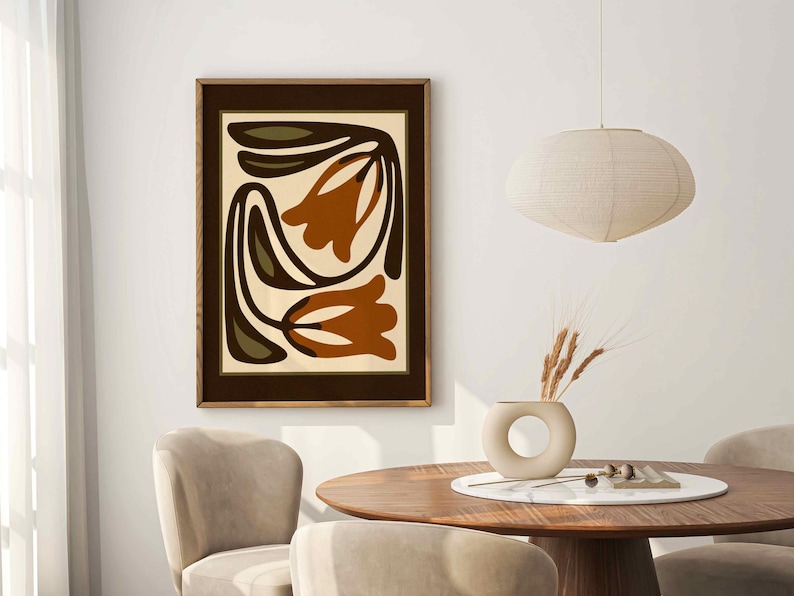 May include: A framed abstract art print featuring stylized orange and brown tulip-like shapes on a cream background. The artwork is in a dark brown frame, displayed above a wooden table with chairs. A beige vase with wheat and a pendant light are also visible.