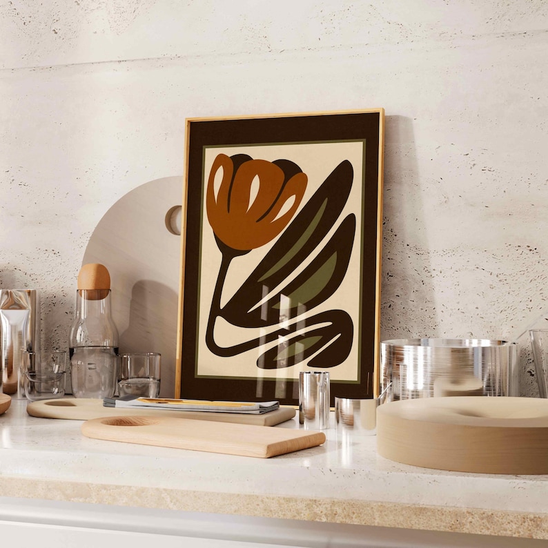 K&ouml;nnte beinhalten: Ein gerahmter Kunstdruck mit einer stilisierten Blume in Braun-, Gr&uuml;n- und Cremet&ouml;nen. Das Kunstwerk befindet sich in einem dunkelbraunen Rahmen und wird auf einer hellen Arbeitsplatte pr&auml;sentiert. Ein modernes Kunstwerk.