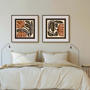 May include: Two framed abstract art prints with brown, olive green, and cream-colored floral designs. The prints are displayed above a bed with white bedding. The frames are dark brown, and the art has a modern, minimalist style.