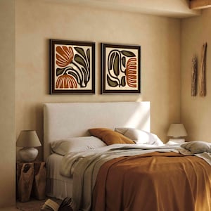 May include: Two framed abstract floral art prints in brown and orange tones hang above a bed with a white headboard. The bed has a rust-coloured blanket and pillows. A wooden side table with a lamp and an open book are visible.