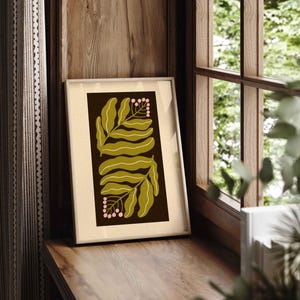 Pu&ograve; includere: Una stampa artistica botanica incorniciata con foglie verde oliva e bacche rosa su sfondo marrone scuro. L'opera d'arte &egrave; in una cornice color crema e poggia su una superficie di legno vicino a una finestra.