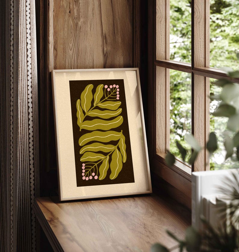 Pu&ograve; includere: Una stampa artistica botanica incorniciata con foglie verde oliva e bacche rosa su sfondo marrone scuro. L'opera d'arte &egrave; in una cornice color crema e poggia su una superficie di legno vicino a una finestra.