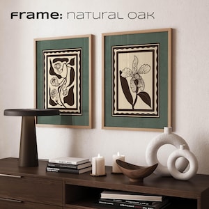 Op de afbeelding: Twee ingelijste botanische kunstprints met natuurlijke eikenhouten frames. De prints hebben een donkergroene achtergrond met een cr&egrave;mekleurig midden en een zwart-cr&egrave;mekleurig bloemmotief. De tekst "Frame: natural oak" staat bovenaan. De prints worden tentoongesteld op een donkerbruin dressoir met decoratieve items.