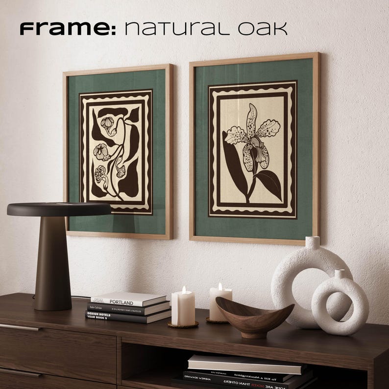 Op de afbeelding: Twee ingelijste botanische kunstprints met natuurlijke eikenhouten frames. De prints hebben een donkergroene achtergrond met een cr&egrave;mekleurig midden en een zwart-cr&egrave;mekleurig bloemmotief. De tekst "Frame: natural oak" staat bovenaan. De prints worden tentoongesteld op een donkerbruin dressoir met decoratieve items.