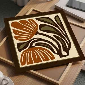 May include: A framed art print featuring a stylized floral design in shades of brown, tan, and olive green. The artwork is set against a cream background and framed in a dark brown border. The print is displayed on a wooden surface.
