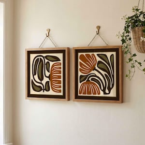 May include: Two framed floral art prints. Each print features a brown and olive green floral design on a cream background. The frames are light brown wood, and the prints hang from brass hooks with chains.