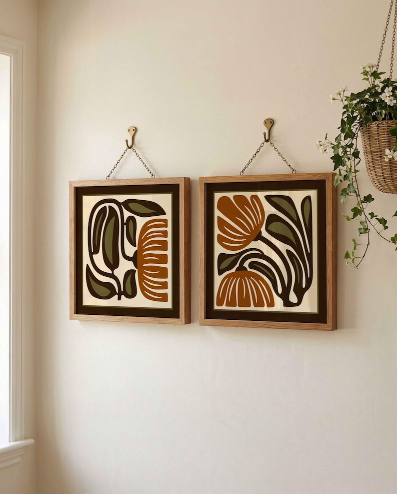 May include: Two framed floral art prints. Each print features a brown and olive green floral design on a cream background. The frames are light brown wood, and the prints hang from brass hooks with chains.