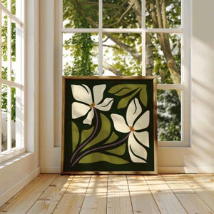 May include: A framed art print depicting two white flowers with brown centers and green leaves on a dark green backdrop. The artwork, in a gold frame, is positioned near a window, illuminated by natural light.