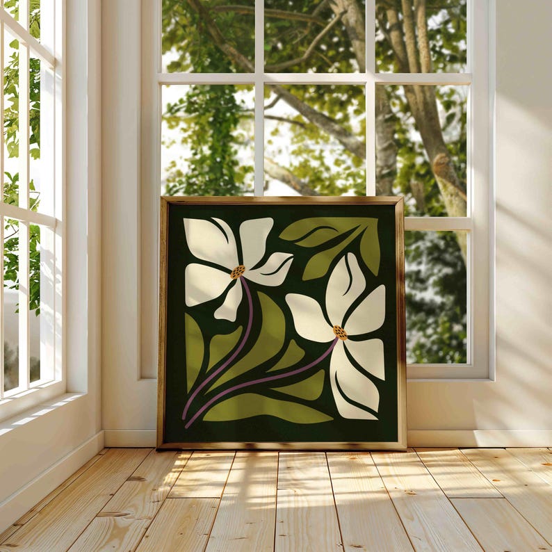 May include: A framed art print depicting two white flowers with brown centers and green leaves on a dark green backdrop. The artwork, in a gold frame, is positioned near a window, illuminated by natural light.