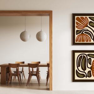 May include: Two framed abstract floral art prints in brown and olive green tones hang on a white wall. The prints feature stylized botanical designs. The room also includes a wooden dining table with chairs and two pendant lights.