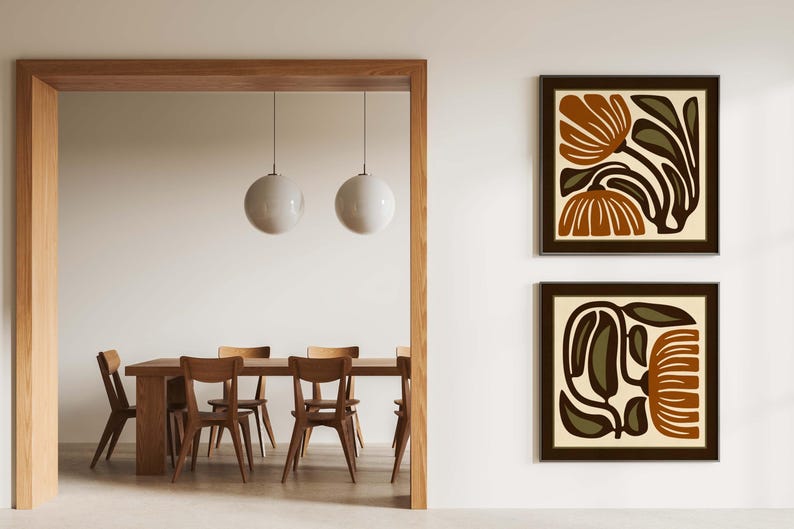 May include: Two framed abstract floral art prints in brown and olive green tones hang on a white wall. The prints feature stylized botanical designs. The room also includes a wooden dining table with chairs and two pendant lights.