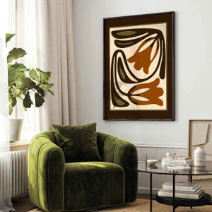 May include: Abstract floral art print in a dark brown frame. The artwork features stylized brown and green floral shapes on a cream background. The framed art hangs above a green velvet armchair in a living room setting.