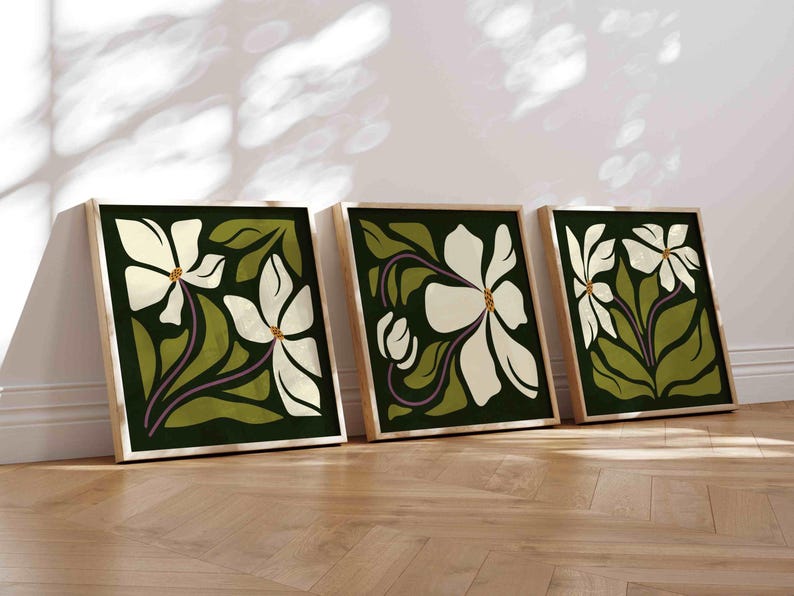 May include: Three framed floral art prints. Each print features a large white flower with a yellow centre and purple stem against a dark green background. The frames are light wood, and the art leans against a white wall.