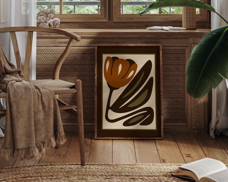 K&ouml;nnte beinhalten: Ein gerahmter Kunstdruck mit einem abstrakten Blumendesign in Braun-, Orange- und Gr&uuml;nt&ouml;nen. Das Kunstwerk steht auf einem Holzboden, gelehnt an einer Wand. Ein Holzstuhl mit einer Decke und einem Teppich befinden sich im Vordergrund.