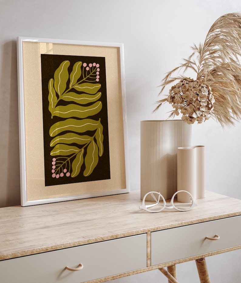 Pu&ograve; includere: Una stampa botanica incorniciata con un bordo bianco. L'opera presenta foglie verde oliva e bacche rosa su uno sfondo scuro. La stampa &egrave; esposta su una scrivania in legno chiaro con vasi decorativi e occhiali.