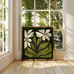 May include: A framed art print depicting two white flowers with brown centres and green leaves on a dark green backdrop. The artwork, in a gold frame, is displayed near a window, showcasing a floral theme.
