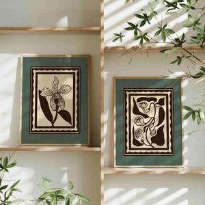 Op de afbeelding: Twee ingelijste botanische kunstprints. Elke print heeft een zwart-wit bloemenontwerp op een cr&egrave;mekleurige achtergrond, geplaatst in een donkergroene lijst. De lijsten worden tentoongesteld op houten planken, met groen in de voorgrond.