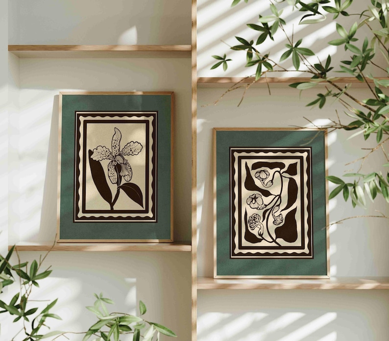 Op de afbeelding: Twee ingelijste botanische kunstprints. Elke print heeft een zwart-wit bloemenontwerp op een cr&egrave;mekleurige achtergrond, geplaatst in een donkergroene lijst. De lijsten worden tentoongesteld op houten planken, met groen in de voorgrond.