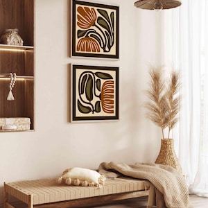 May include: Two framed abstract floral art prints in shades of brown, olive green, and cream hang on a white wall. The artwork is displayed above a wooden bench with a cream-colored pillow and a woven rug. A woven pendant light hangs from the ceiling.