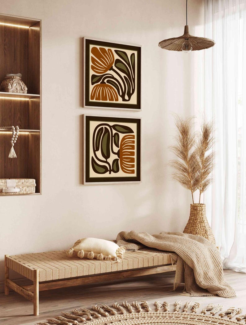 May include: Two framed abstract floral art prints in shades of brown, olive green, and cream hang on a white wall. The artwork is displayed above a wooden bench with a cream-colored pillow and a woven rug. A woven pendant light hangs from the ceiling.