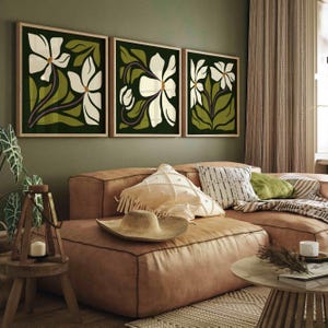 May include: Three framed floral art prints. Each print features a white flower with green leaves and purple stems against a dark green background. The frames are a light wood colour. The artwork is displayed above a brown leather sofa.