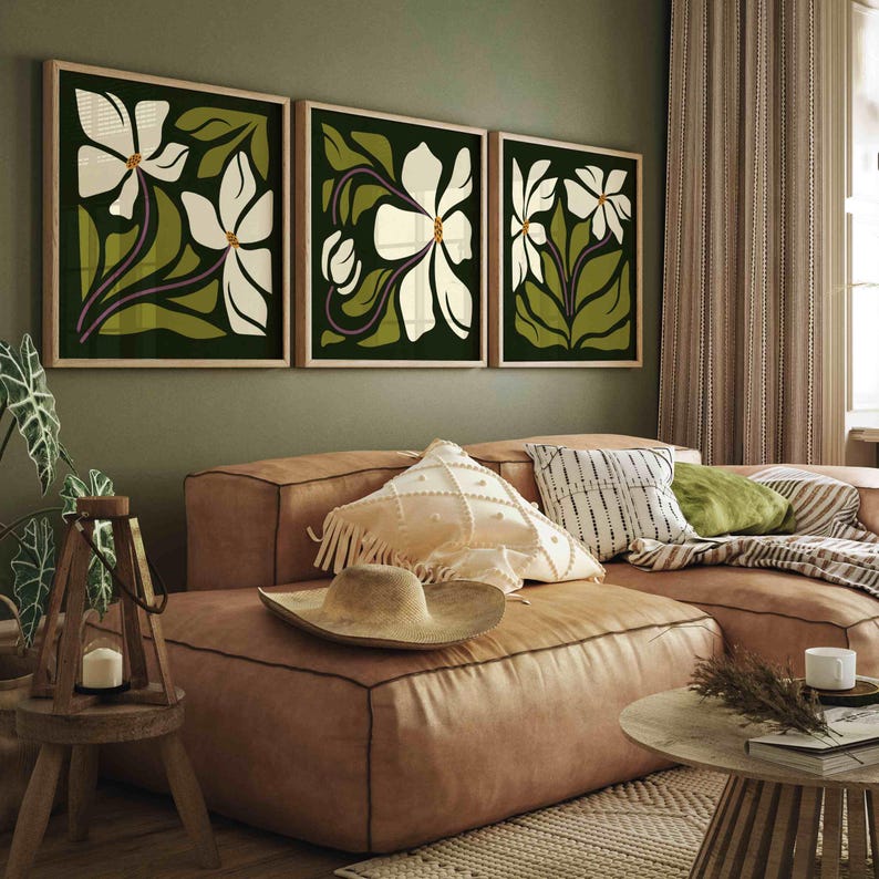 May include: Three framed floral art prints. Each print features a white flower with green leaves and purple stems against a dark green background. The frames are a light wood colour. The artwork is displayed above a brown leather sofa.