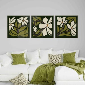 May include: Three framed floral art prints. Each print features a white flower with green leaves and stems against a dark green background. The artwork hangs above a white sofa with green and patterned cushions and a green throw.