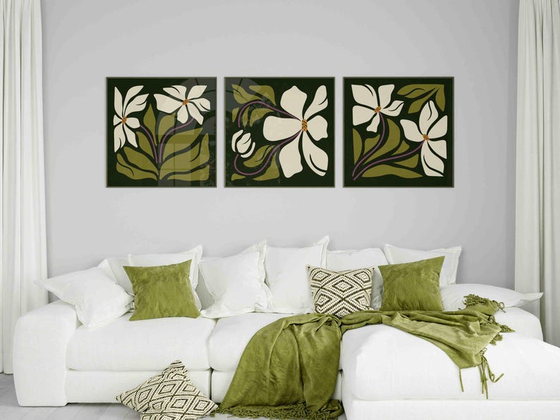 May include: Three framed floral art prints. Each print features a white flower with green leaves and stems against a dark green background. The artwork hangs above a white sofa with green and patterned cushions and a green throw.
