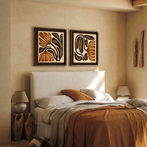 May include: Two framed abstract art prints with brown frames hang above a bed with a white headboard. The bed has a rust-colored blanket and pillows. A wooden side table with a lamp and a book are next to the bed. The room has a neutral color palette.