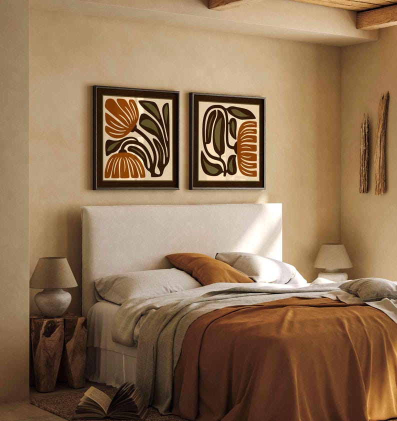 May include: Two framed abstract art prints with brown frames hang above a bed with a white headboard. The bed has a rust-colored blanket and pillows. A wooden side table with a lamp and a book are next to the bed. The room has a neutral color palette.