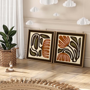 May include: Two framed abstract floral art prints. Each print features a beige background with brown and orange botanical designs. The frames are dark brown. The artwork is displayed on a wooden floor.