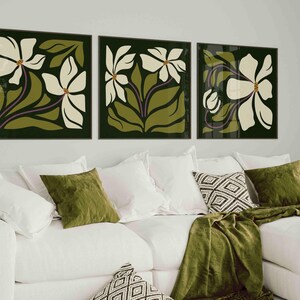 May include: Three framed floral art prints. Each print features a white flower with green leaves and a dark green background. The artwork is displayed above a white sofa with green and patterned cushions and a green throw.