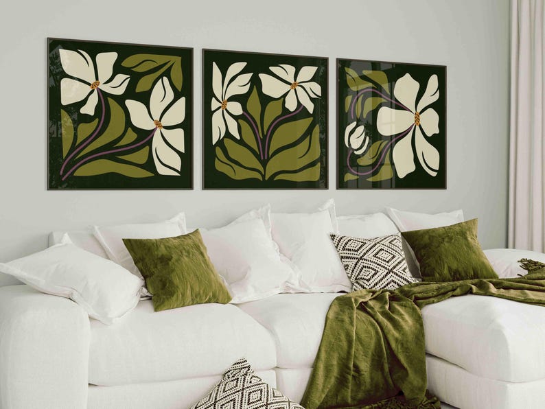 May include: Three framed floral art prints. Each print features a white flower with green leaves and a dark green background. The artwork is displayed above a white sofa with green and patterned cushions and a green throw.